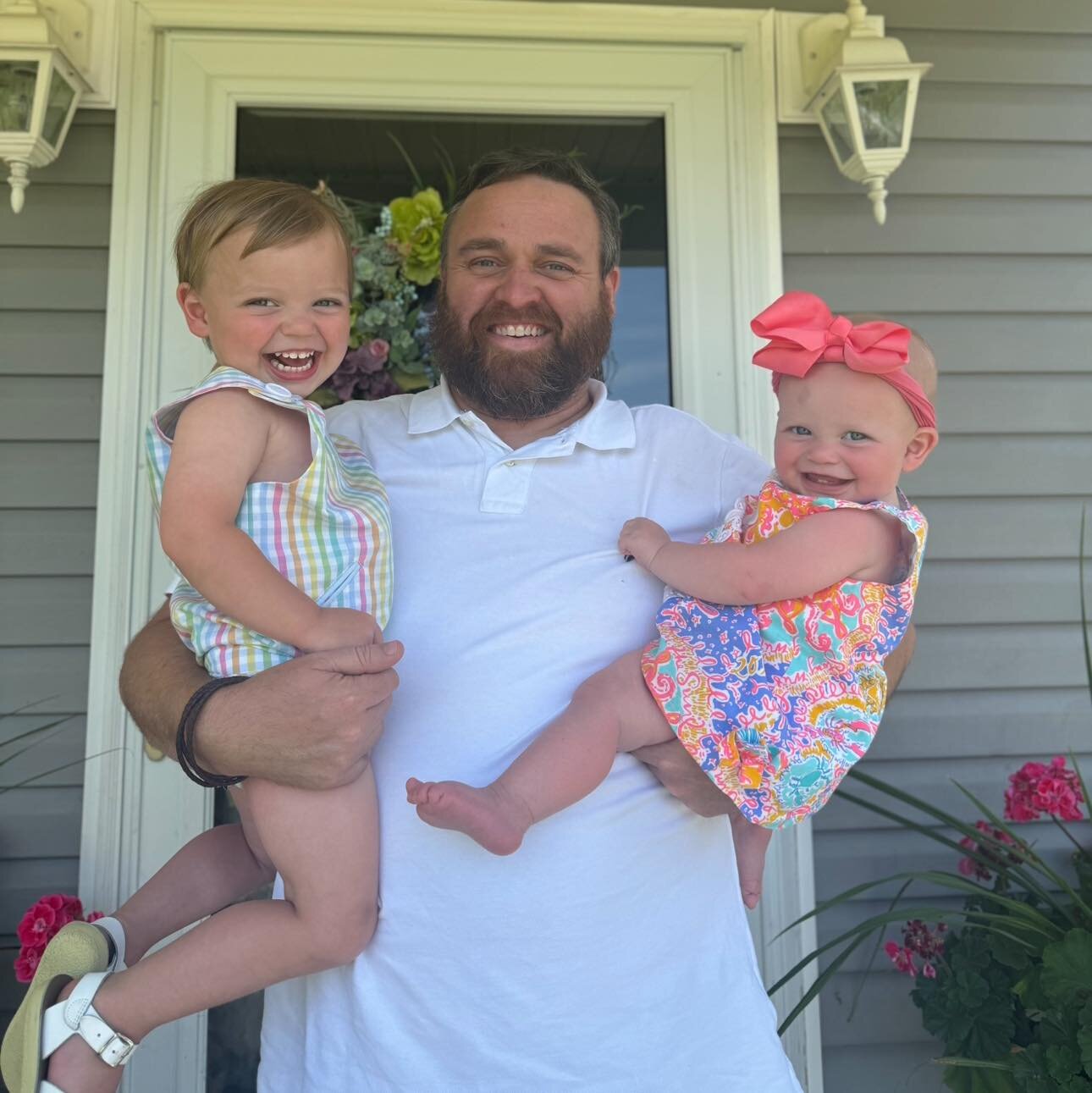 A man with a beard smiles while holding two young children, both smiling, standing outside in front of a house with flowers and a wreath on the door.
