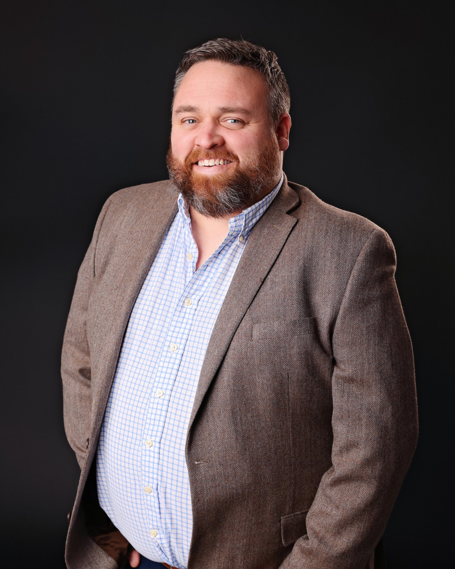 Smiling man with a beard wearing a brown blazer and checkered shirt, standing against a plain dark background.